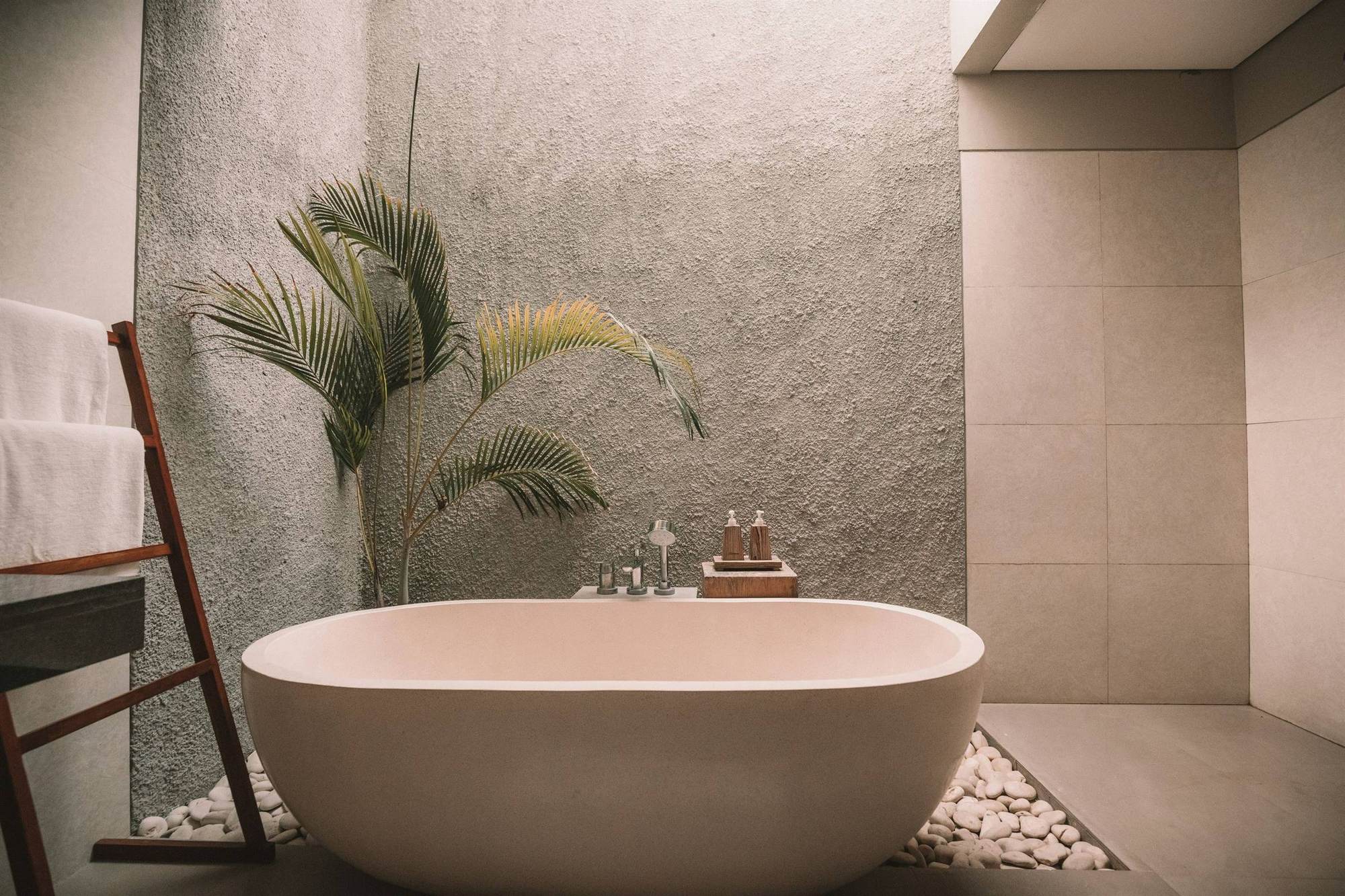 A freestanding stone bath against a textured render wall, a potted palm beside it, white river pebbles set into the floor and a timber towel ladder.