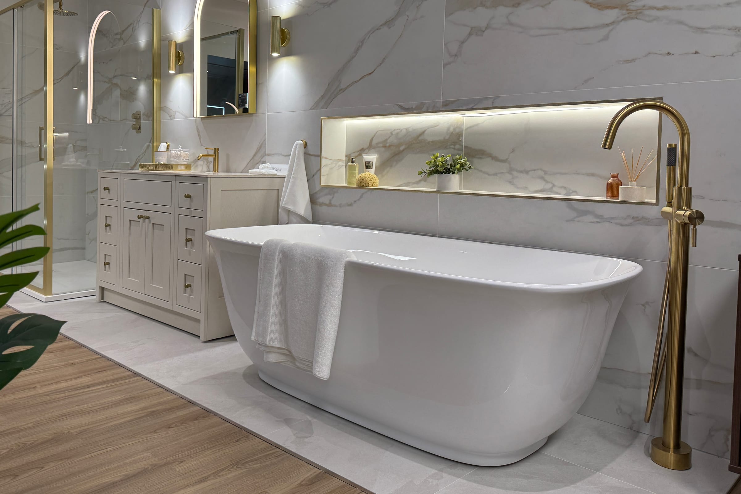 The Poole showroom: a white freestanding bath with a brushed brass floor-standing bath filler, an illuminated recessed niche holding a sponge, a small plant and a reed diffuser, and a cream shaker vanity with brass handles under an arched brass mirror, against Calacatta marble-effect porcelain.