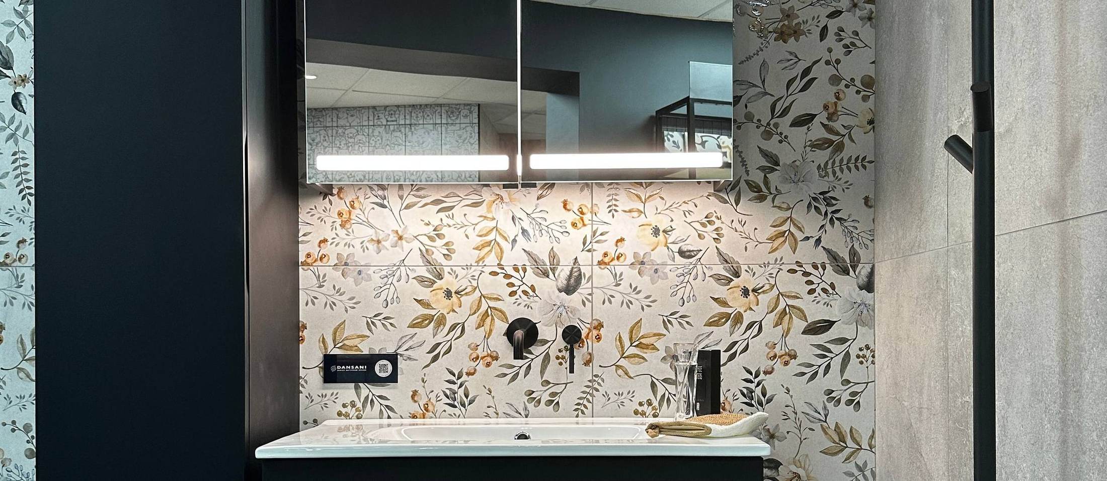 A vanity bay in the Southbourne showroom: a white basin on a dark vanity against a floral patterned tile wall, with black wall-mounted taps, a backlit mirrored cabinet above and grey stone-effect tiling alongside.