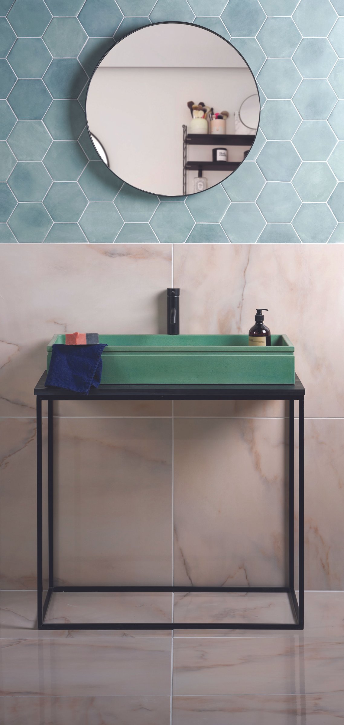 A green concrete trough basin on a slim black metal washstand, a round black-framed mirror above, set against blue-green hexagonal tiles and marble-effect tiling.