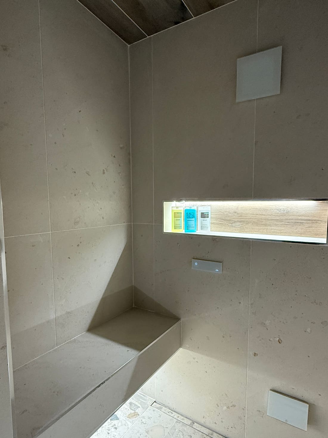 Inside the steam shower display at the Poole showroom. A built-in corner bench in stone-effect tile, a lit recess lined in wood-effect tile holding three bottles, two flush white square panels on the walls and a wood-effect tiled ceiling
