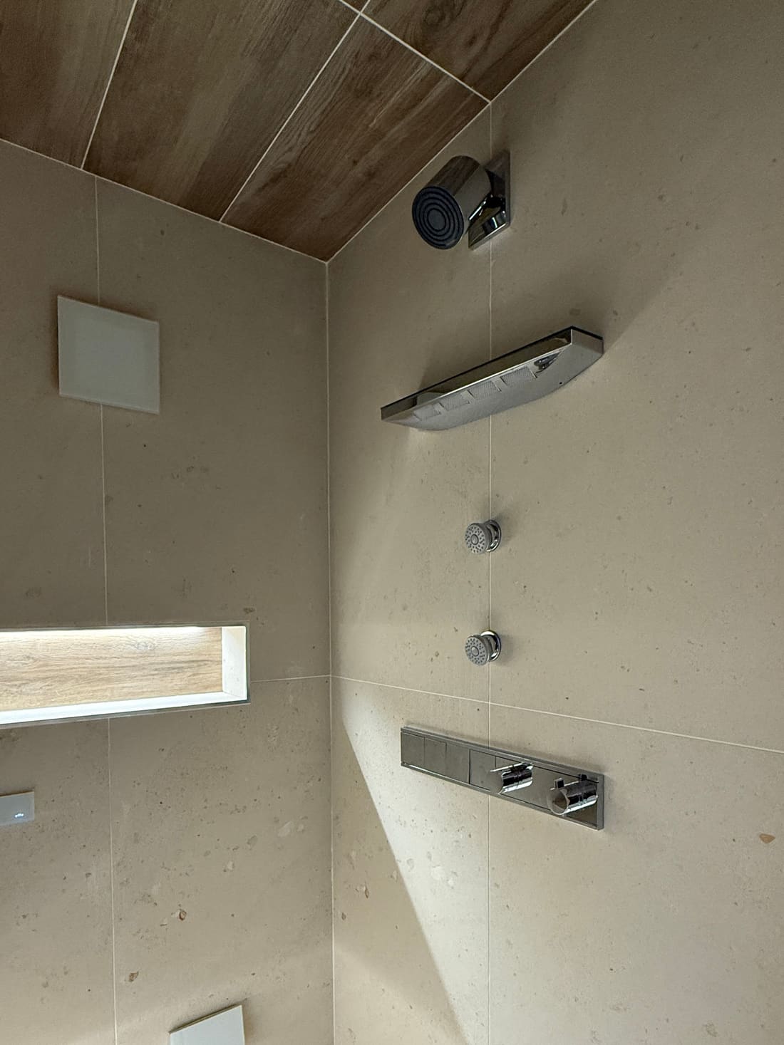 Inside the steam shower display at the Poole showroom. A rectangular chrome shower head and a round chrome and black fitting are mounted on a stone-effect tiled wall, with two round body jets, a chrome control bar, a flush white square panel, a lit recessed shelf and a ceiling clad in wood-effect tile