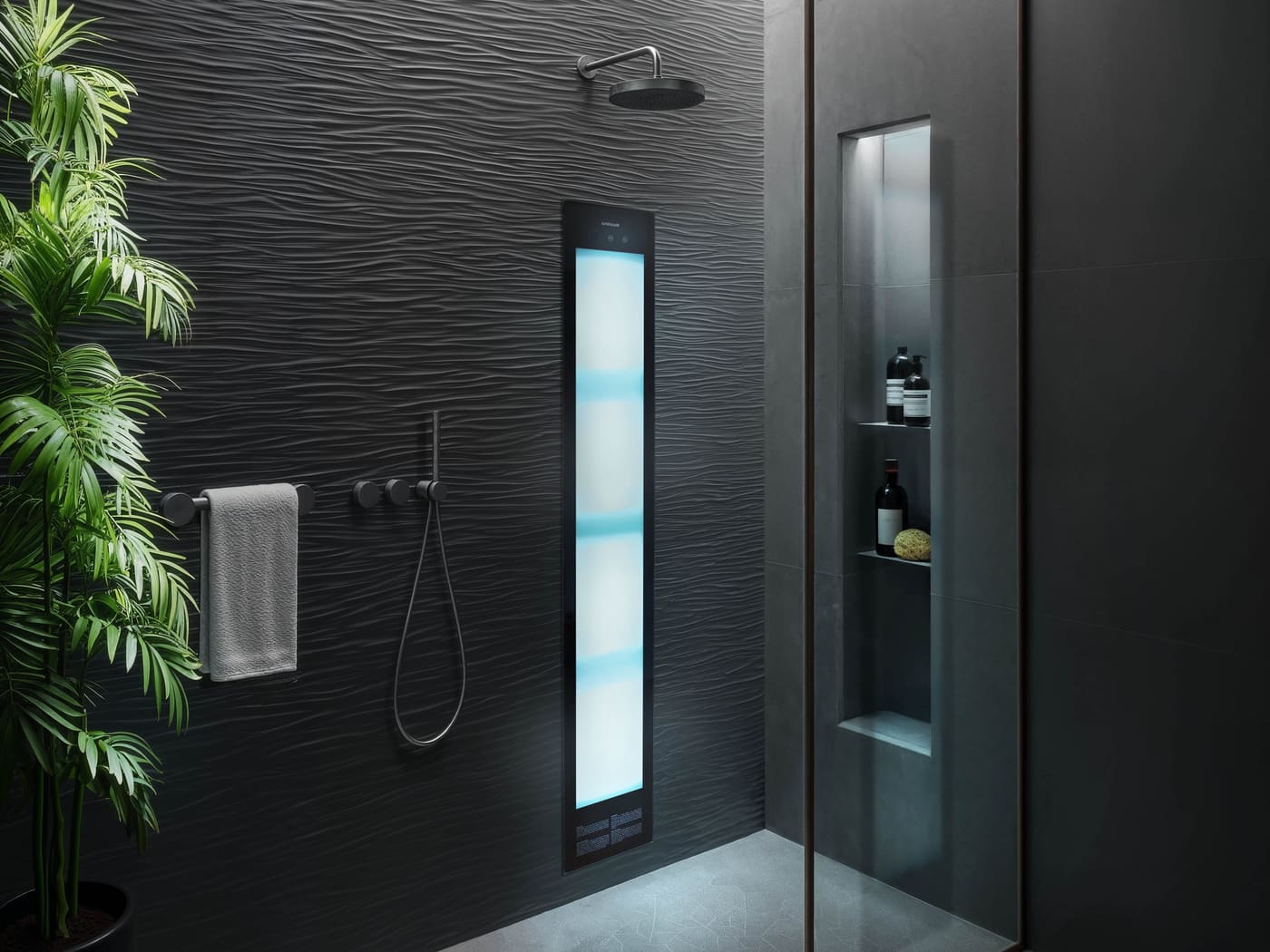 A dark shower lined in black wave-textured tile. A Sunshower panel glows cool blue and white beside matt black controls, a hand shower and a grey towel on a rail. A palm stands at the left and a lit niche behind the glass holds bottles