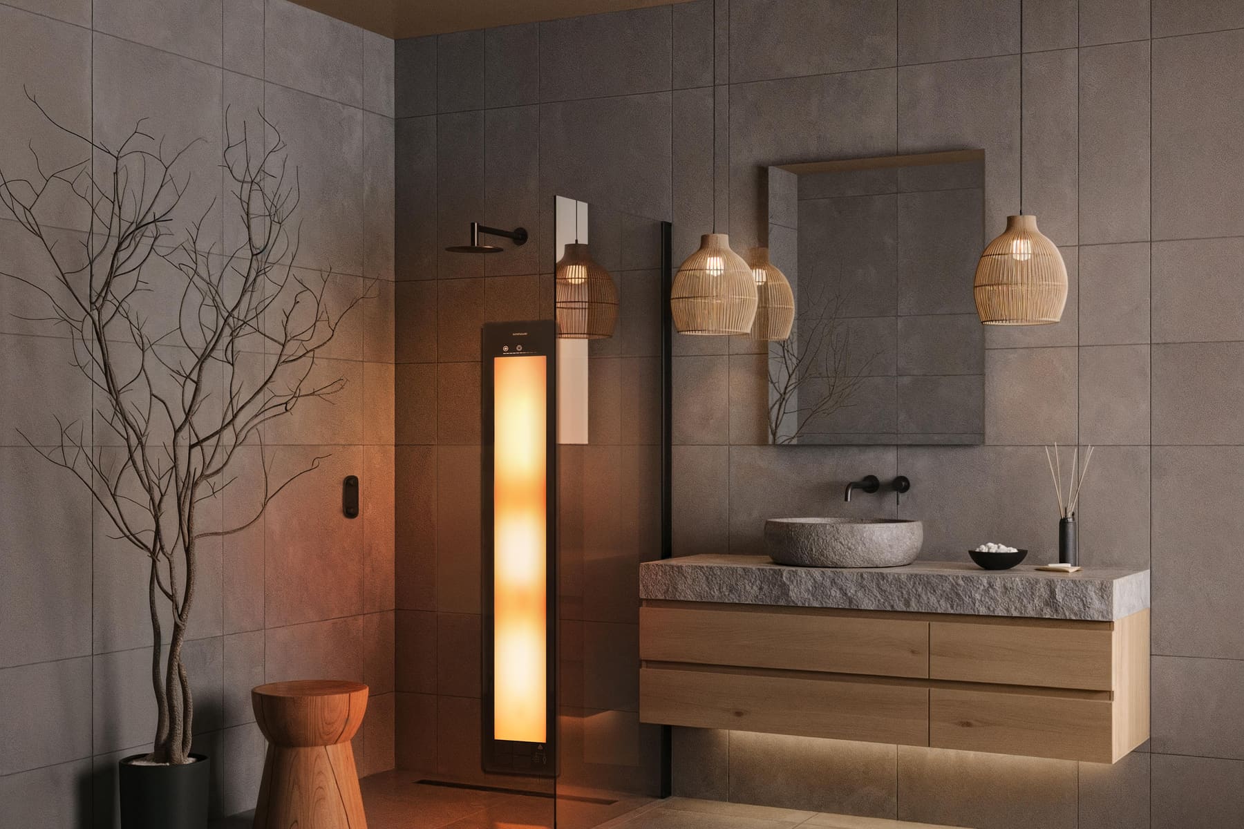 A bathroom lined in large grey stone-effect tiles. A tall Sunshower panel glows warm amber inside a walk-in shower behind a glass screen. To the right an oak vanity carries a rough stone basin, with three woven pendant lights and a mirror above, and a bare-branch tree in a black pot at the left