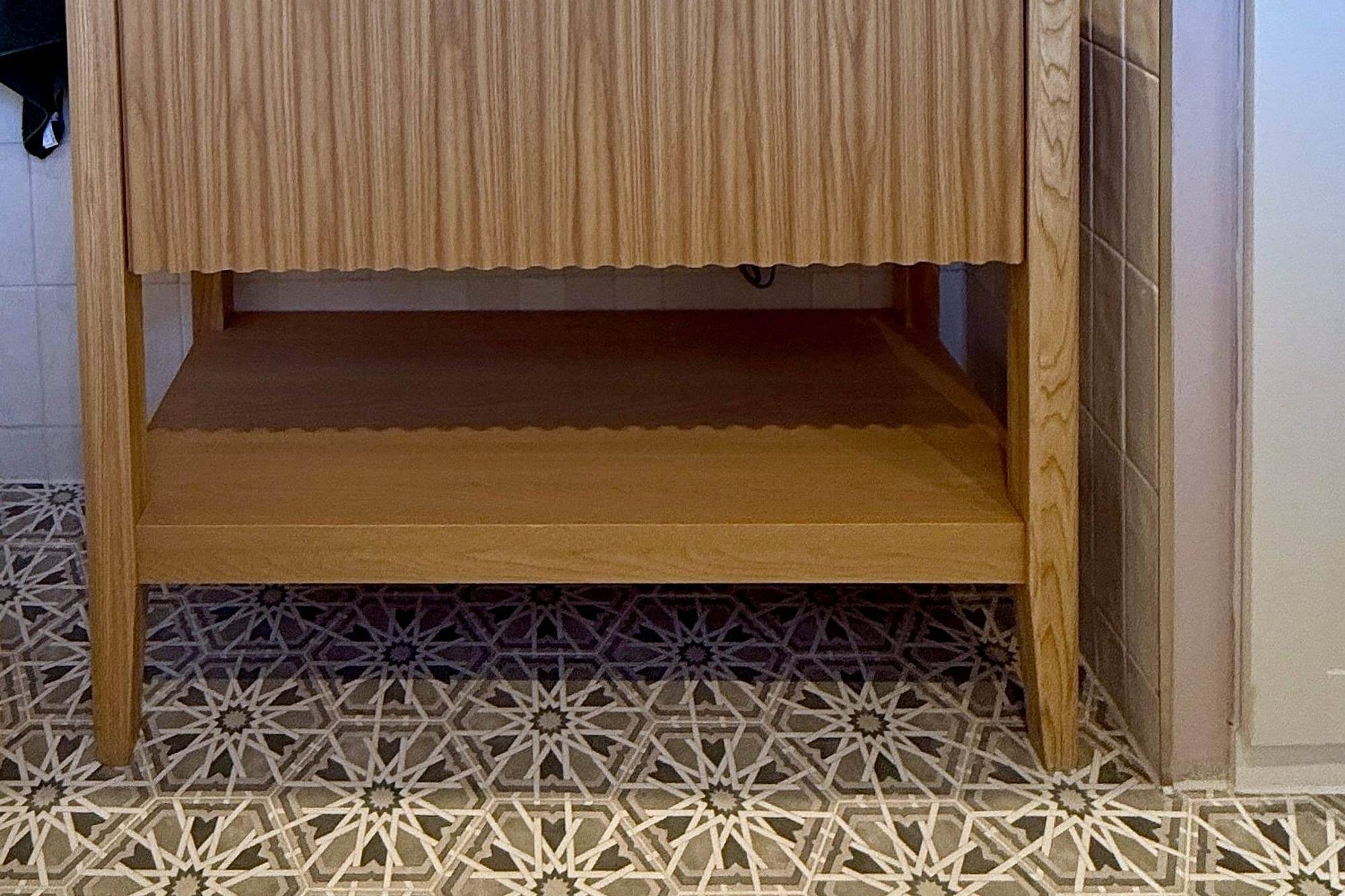 A freestanding light oak washstand seen head on, its finely fluted drawer front above an open lower shelf and square legs, standing on a bold star-pattern floor tile that runs wall to wall, with pale handmade-look tile on the wall behind.