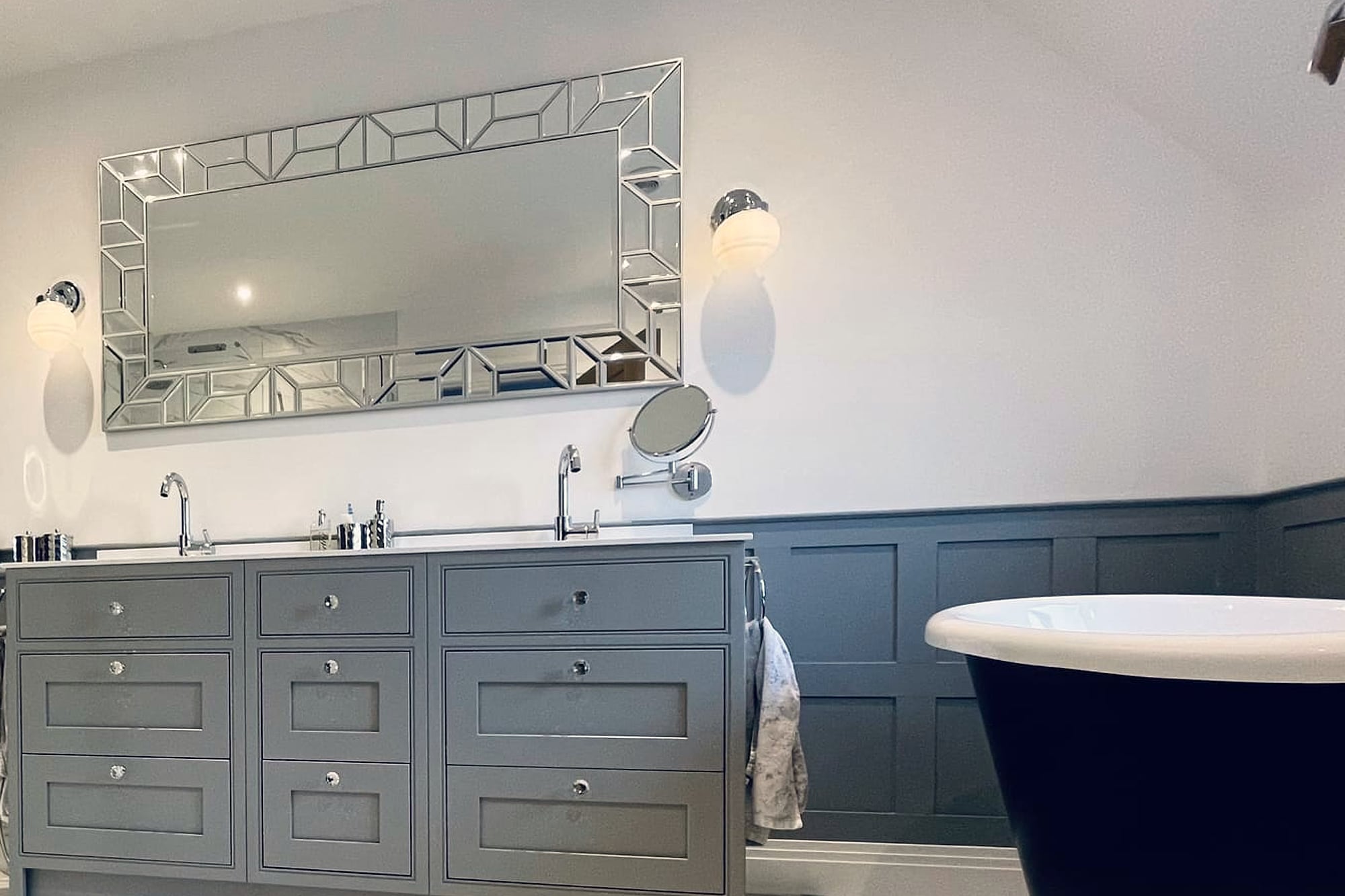A bathroom in grey framed panelling: a nine-drawer double vanity with a white top and two chrome mixers, under a rectangular mirror in a stepped bevelled border with a wall light either side and an extending magnifying mirror beside it, and at the right the white rim and dark outer skin of the boat bath, above a large-format marble-effect porcelain floor.