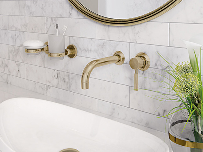 Brushed brass wall-mounted taps on white marble-effect tiling, with the lower edge of a round brass-framed mirror above.